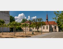 Ein kleiner Platz mit gepflastertem Weg und jungen Bäumen führt zu einer hellen, historischen Kirche mit roten Ziegeldächern und einem spitzen Turm. Im Hintergrund sind Gebäude, Bäume und ein blauer Himmel mit weißen Wolken zu sehen.