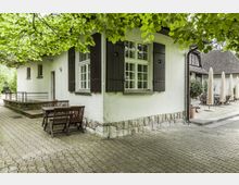 Ein weißes Gebäude mit dunklen Holzfensterläden und einem Steinsockel, umgeben von einem gepflasterten Hofbereich. Im Vordergrund stehen ein Holztisch mit Stühlen unter einem Baum, im Hintergrund sind Sonnenschirme und weitere Sitzgruppen zu sehen.