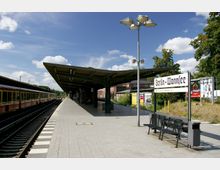 Bahnhof mit überdachtem Bahnsteig und einem Schild mit der Aufschrift „Berlin-Wannsee“. Links befindet sich ein abgestellter Zug, rechts Werbeplakate und Sitzbänke, umgeben von Bäumen und blauem Himmel.