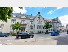 Straßenansicht eines historischen Gebäudes mit heller Fassade, Satteldach und Fenstern im traditionellen Stil. Im Vordergrund stehen geparkte und fahrende Autos sowie ein Gehweg mit Bäumen und einem Radwegschild.