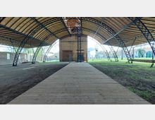 Offene, überdachte Struktur mit einem gebogenen Holz- und Metallgerüst, das auf beiden Seiten von eisernen Bögen gestützt wird. Im Hintergrund steht ein Backsteingebäude, umgeben von einer Grünfläche mit industriellen Objekten und historischen Elementen.