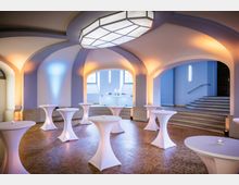 Eleganter Veranstaltungsraum mit gewölbten Decken, beleuchteten Stehtischen und warmem indirektem Licht. Im Hintergrund führt eine Treppe zu einem erhöhten Bereich mit einem weißen Tresen und Fenstern.