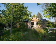 Backstein-Häuschen mit weißen Fensterläden in einem grünen Garten mit Rasenfläche, bunten Blumenbeeten und hohen Bäumen; weiße Gartenmöbel stehen verteilt im Schatten und in der Sonne.