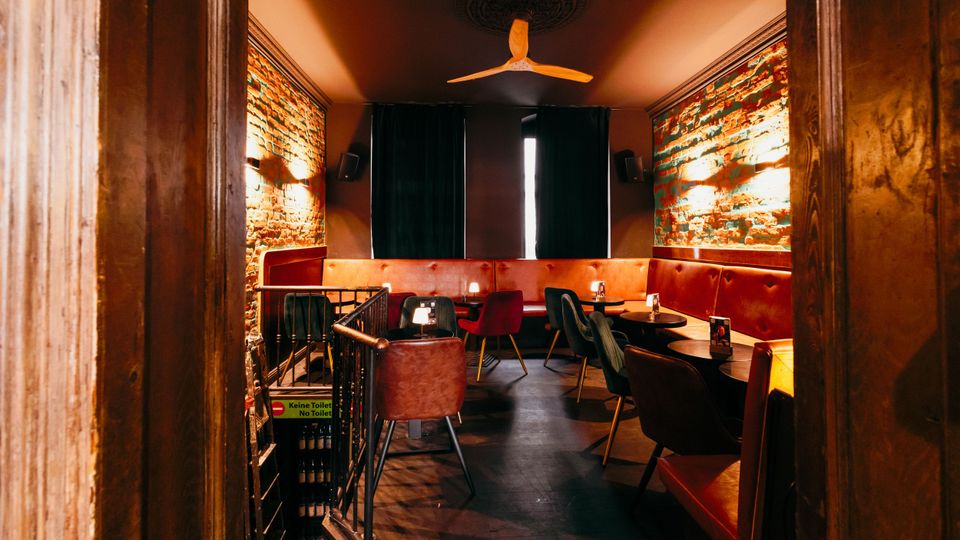 Blick in einen schmalen Bar- oder Loungebereich mit freigelegten Backsteinwänden, roter gepolsterter Sitzbank und kleinen runden Tischen mit Stühlen. An der Decke hängt ein Ventilator, am Ende des Raums sind dunkle Vorhänge, seitliche Wandleuchten und ein Holzrahmen am Eingang zu sehen.