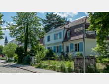 Straßenecke mit gepflegtem Wohnhaus im traditionellen Stil, hellblauen Fensterläden und rotem Ziegeldach, umgeben von üppiger Vegetation; Straßenschilder weisen auf „Kirchblick“ und „Salzachstraße“ hin. Im Hintergrund ragt ein Kirchturm zwischen den Bäumen hervor.