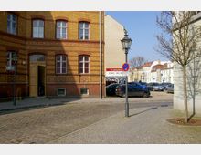 Straßenszene mit einem historischen Gebäude aus gelben und roten Backsteinen mit hohen Fenstern und einer geöffneten Eingangstür. Im Vordergrund befindet sich eine gepflasterte Straße, ein Straßenschild mit Parkplatzhinweis und ein kahler Baum, im Hintergrund sind ein Parkplatz und weitere Häuser zu sehen.