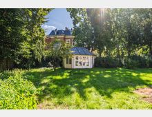 Grüne Gartenanlage mit großer Rasenfläche und hohen Laubbäumen; in der Mitte steht ein heller Pavillon mit vielen Fenstern und achteckigem Dach. Im Hintergrund ist ein mehrstöckiges Backsteingebäude mit Schieferdach teilweise durch das Blattwerk zu sehen.