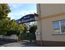 Eingang zu einem Biergarten mit einem Schild mit der Aufschrift „Worrick's Biergarten“ über einem grünen Tor. Im Hintergrund sind Bäume, Pflanzen und ein Teil der Außenbestuhlung sichtbar, daneben eine beige Hauswand mit einem Fenster.