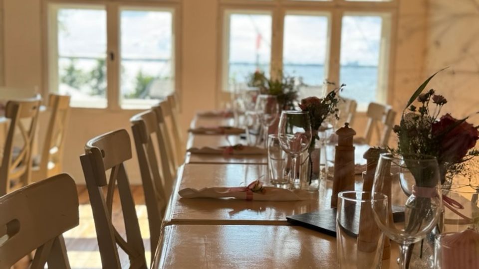 Langer, festlich gedeckter Holztisch in einem Raum mit weiß gestrichenen Wänden, großen Sprossenfenstern und einer Decke mit sichtbaren Holzbalken. Im Hintergrund ist durch die Fenster eine Aussicht auf eine Landschaft zu erkennen.