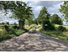 Ein schmaler, unbefestigter Weg führt zwischen zwei bewachsenen Torpfeilern in eine ländliche Landschaft mit Bäumen, Wiesen und Zaunanlagen. Der Weg ist von grüner Vegetation und Schatten durch die Bäume gesäumt, während der Himmel teils wolkig ist.