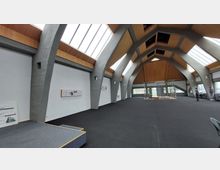 Innenraum eines modernen Gebäudes mit großen, bogenförmigen Betonstützen, einer Holzdecke und breiten Oberlichtern, die viel Tageslicht einlassen. Der Raum hat einen schwarzen Teppichboden, eine kleine Bühne links und eine Baustelle mit Gerüst in der Mitte.