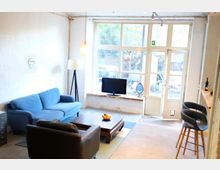 Helles Wohnzimmer mit großen Fenstern und einer Glastür, die auf einen begrünten Außenbereich führt. Der Raum ist mit einem blauen Sofa, einem Ledersessel, einem Holz-Couchtisch und einer Küchenzeile mit Barhockern ausgestattet; ein Fernseher steht auf der Fensterbank.