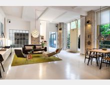 Helles Wohnzimmer in modernem Loft-Stil mit hohen Decken, freiliegenden Ziegelwänden und großen Fenstern. Der Raum ist eingerichtet mit einem Sofa, Sesseln, einem Couchtisch auf grünem Teppich, Designerlampen und einem Essbereich mit Holztisch und Stühlen.