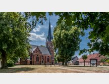 Backsteinkirche mit hohem Spitzdach-Turm in einem kleinen Dorf, umgeben von großen Bäumen und einem Rasenplatz; im Hintergrund stehen Wohnhäuser.