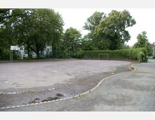 Ein leerer, asphaltierter Sportplatz mit einem Basketballkorb auf der linken Seite und einem kleinen Fußballtor im Hintergrund. Der Platz ist von Bäumen und Büschen umgeben, im Hintergrund sind Wohngebäude zu sehen.