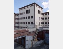 Mehrstöckiges Gebäude mit hellen Wänden und roten Backsteinkanten, umgeben von einer Ziegelmauer. Im Vordergrund ist eine offene Metalltür und eine Außentreppe sichtbar.