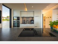 Moderne Küche mit hellen Einbauschränken, integriertem Backofen, Mikrowelle und Kühlschrank. Eine Arbeitsfläche enthält ein schwarzes Kochfeld, im Hintergrund ein Fenster mit Blick auf ein Gewässer und angrenzende Häuser.