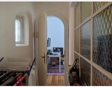 Blick durch einen schmalen Flur mit einem kleinen Fenster und einer gläsernen Trennwand mit rundem Muster zu einem angrenzenden Raum. Der Raum enthält einen Schreibtisch mit einem Monitor, und im Flur steht ein Wäscheständer.