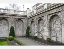 Hof mit gepflastertem Weg, flankiert von einer mit Kletterpflanzen bewachsenen Steinmauer mit mehreren Arkaden. Im Vordergrund sind Hecken und Sträucher zu sehen, die formell geschnitten sind.