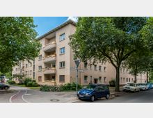 Mehrstöckiges Wohngebäude in beige-cremefarbener Fassade mit Balkonen, umgeben von Bäumen und gepflegten Grünflächen. Im Vordergrund befindet sich eine Straße mit parkenden Autos sowie ein Straßenschild, das die Kreuzung "Bismarckstraße" und "Friedrichstraße" markiert.