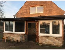 Ein renovierungsbedürftiges Haus mit verputzter Außenwand und einem Anbau mit großen weißen Fenstern und Backsteinmauerwerk. Im Vordergrund ein kahler Gartenbereich mit einem Holzstuhl und kleinen Pflanzen.