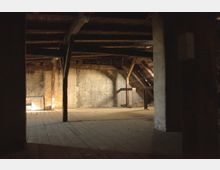 Ein leerer, alter Dachboden mit Holzbalken, Holzfußboden und freiliegenden, schrägen Dachstrukturen. Tageslicht fällt durch Spalten in die dunkle, staubige Umgebung.