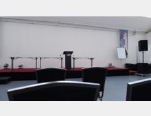 Ein leerer Veranstaltungsraum mit einer Bühne. Auf der Bühne stehen ein Rednerpult mit der Aufschrift „Forum“, zwei leere Tischgestelle, ein Flipchart, schwarze Lautsprecher und eine Wanddekoration. Im Vordergrund sind schwarze Stuhllehnen sichtbar.