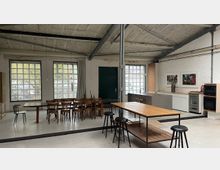 Großer, offener Atelier- oder Werkstattraum mit hoher Betondecke und Stahlträgern, großen Glasbausteinfenstern und hellem Boden. Mehrere Holztische und Stühle stehen im Raum, rechts eine kleine Küchenzeile und Wandbilder.