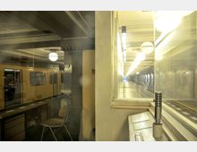 Innenansicht einer U-Bahn-Station mit gelbem Zug auf der linken Seite, einer leeren Wartehalle im Hintergrund und einem Schalterraum mit Glasfenster sowie einer Tischplatte, Mikrofon und Stuhl im Vordergrund. Die Decke ist mit Zierelementen versehen, und runde Leuchten hängen entlang des Bahnsteigs.