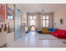 Wohnzimmer mit hellem Holzboden, hohen Decken und weißen Vorhängen, die große Fenster und eine geöffnete Balkontür einrahmen. Farbig gestaltete Stoffsofas in Rot, Blau und Gelb sowie eine cremefarbene Wand mit dekorativen Bildern prägen den Raum.