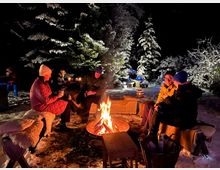 Vier Personen sitzen bei Nacht um ein Lagerfeuer in einem schneebedeckten Wald. Im Hintergrund sind mit Schnee bedeckte Bäume, Laternen und warme Beleuchtung sichtbar.