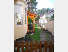 Gartenansicht mit einem gelb gestrichenen Haus auf der linken Seite, einer kleinen überdachten Terrasse aus Holz und angrenzendem Rasen. Rechts ist ein weißer Lattenzaun zu sehen, dahinter befinden sich weitere Häuser, umgeben von Bäumen.