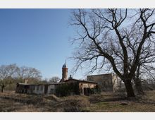 Verlassen wirkendes Gelände mit niedrigem Backstein- und Putzgebäude, dahinter ein hoher runder Turm mit Kuppeldach. Im Vordergrund stehen ein großer kahler Baum und trockenes, überwuchertes Gras unter blauem Himmel.