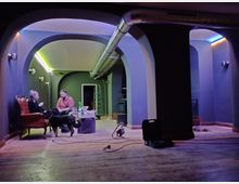 Ein Raum mit dunklen, gewölbten Wänden, indirekter Beleuchtung in verschiedenen Farben und einem hölzernen Boden. Zwei Personen sitzen auf Sesseln neben einem niedrigen Tisch, während ein Hund daneben steht; im Hintergrund sind eine Leiter, eine geöffnete Tür und industrielle Rohrleitungen sichtbar.