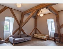 Wohnraum mit freiliegenden Holzbalken und großen, weiß gerahmten Fenstern. Der Raum ist mit einem grauen Sofa, einem geblümten Sofa und einem Holzstuhl eingerichtet, mit hellem Holzboden und neutraler Wandfarbe.