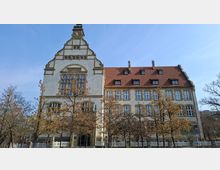 Historisches mehrstöckiges Gebäude mit heller Fassade, großen Sprossenfenstern und hohem geschwungenem Giebel mit Uhrturm, daneben ein langer Flügel mit rotem Ziegeldach und Gauben. Davor stehen mehrere laublose Bäume entlang eines Zauns unter blauem Himmel.