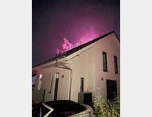 Ein modernes weißes Wohnhaus bei Nacht, mit Feuerwerk in pinken und violetten Farben, das den Himmel über dem Dach erhellt. Im Vordergrund sind Teile eines schwarzen Autos und ein Gartenzaun sichtbar.