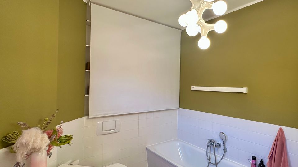 Modernes Badezimmer mit olivgrün gestrichenen Wänden und weiß gefliester Badewanne an der rechten Seite, dazu Waschmaschine und WC links. An der Decke hängt eine kugelförmige Mehrfachleuchte, über dem WC ist ein heruntergezogenes weißes Rollo, der Boden ist hell gefliest.