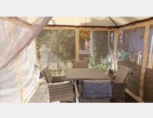 Überdachte Gartenterrasse in einem Zeltpavillon mit lichtdurchlässigen Seitenteilen, ausgestattet mit einem rechteckigen Tisch und mehreren Rattan-Sesseln. Im Hintergrund sind ein umzäunter Garten mit Bäumen und Sträuchern sowie angrenzende Wohngebäude zu sehen.
