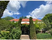 Zweistöckiges Wohnhaus mit weißer Fassade und rotem Ziegeldach, umgeben von üppigem Grün, darunter hohe Sträucher und gepflegte Bäume. Im Vordergrund eine überdachte Terrasse mit Stufen, die von Kletterpflanzen eingerahmt ist, unter einem sonnigen Himmel mit weißen Wolken.