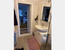 Badezimmer mit weiß gefliesten Wänden, Waschbecken auf einem weißen Unterschrank, einem Spiegel mit seitlicher Ablage und einem Handtuchhalter. Im Hintergrund ist durch eine offene Tür ein Schlafzimmer mit einem Bett und einem Fenster zu sehen.