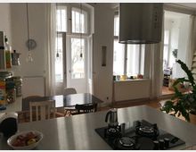 Helle Wohnküche mit hohen Decken und großen, weißen Doppelfenstern. Im Vordergrund befindet sich eine Kücheninsel mit einem Gaskochfeld, im Hintergrund ein Esstisch mit Stühlen, ein Heizkörper und dekorative Pflanzen.