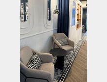 Zwei beige Polstersessel mit gemusterten Kissen stehen neben einem kleinen schwarzen Tisch auf einem gemusterten Fliesenboden. Im Hintergrund befinden sich eine weiße Wand mit integrierten Spiegeln, dunkle Vorhänge und ein Regal mit dekorativen Elementen.