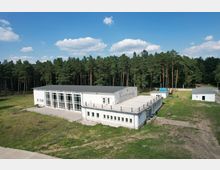 Ein einstöckiges, weißes Gebäude mit großen Glasfenstern und Flachdach, umgeben von einer Wiese und einem angrenzenden Kiefernwald. Im Hintergrund sind weitere kleinere Gebäude und ein blauer Himmel mit wenigen Wolken sichtbar.