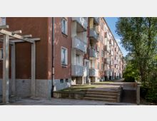 Mehrstöckiges Wohngebäude mit rötlicher Fassade, weißen Balkonen und verputztem Sockelbereich, umgeben von Gehwegen, kleinen Grünflächen und Bäumen. Eine niedrige Treppe führt zu einem Weg entlang der Gebäudeseite.