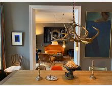 Blick von einem Essbereich mit langem Holztisch und Kerzenhaltern durch eine breite, weiß gerahmte Türöffnung in ein angrenzendes Wohnzimmer. Im Wohnzimmer stehen ein orangefarbenes Sofa, dunkle Sessel und ein Couchtisch auf einem gemusterten Teppich; im Vordergrund hängt eine Geweihleuchte mit freiliegenden Glühbirnen, und an den grauen Wänden hängen gerahmte Bilder.