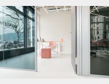 Blick in ein modernes Büro mit großen Glaswänden, hellem Fußboden und minimalistischer Einrichtung. Zu sehen ist ein Schreibtisch mit einem Stuhl und einem rosa Pouf, dahinter eine Fensterfront mit Blick auf parkende Fahrzeuge und Bäume.