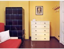 Ein kleiner Raum mit gelber Wand, einem schwarz gefliesten Kachelofen links, einem weißen Sofa mit rotem Bezug und Kissen im Vordergrund. Rechts stehen eine weiße Kommode mit schwarzen Knöpfen, ein gerahmtes Porträt an der Wand und eine hölzerne Garderobenleiste mit roten Knöpfen.