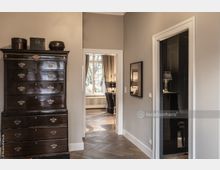 Innenansicht eines eleganten Wohnbereichs mit Parkettboden. Im Vordergrund steht eine dunkle Kommode mit mehreren Schubladen, dahinter öffnet sich ein Durchgang zu einem Raum mit einem Fenster, langen Vorhängen und einem Schreibtischstuhl.