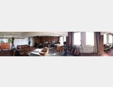 Panoramablick in einen loftartigen Raum mit abgenutzten, teils unverputzten Wänden, großen Fenstern und Teppichboden. Zu sehen sind Sofa- und Sitzecke, ein Schreibtisch sowie Musikstudio-Equipment und mehrere an Ständern stehende Gitarren; rechts schließen Regale und ein Nebenbereich an.